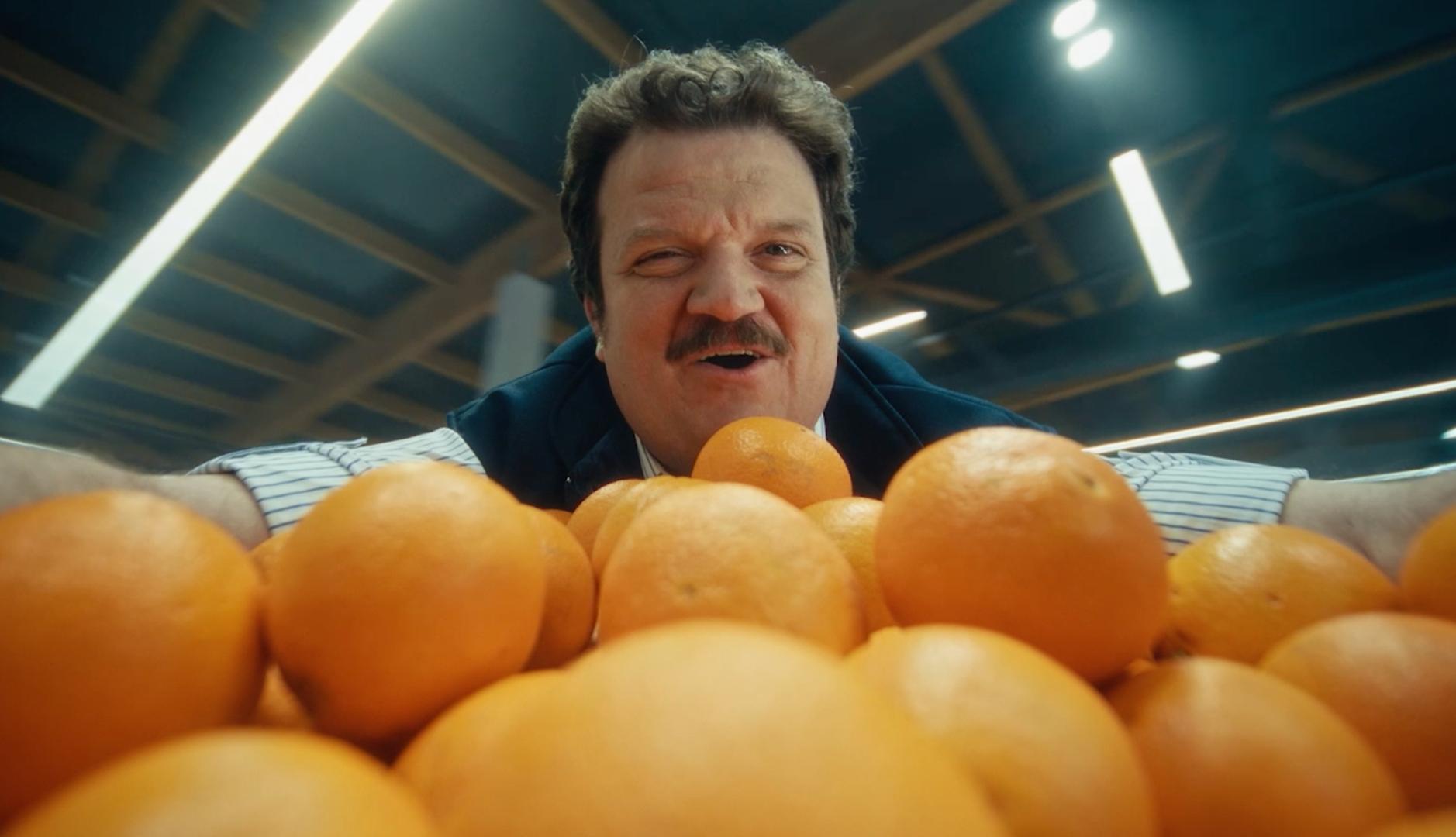 Un cajero canta apoyándose en la estantería de las naranjas de un supermercado