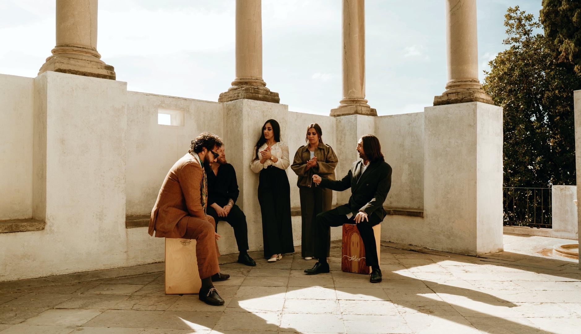 Los artistas del tema "Sosegá" juntos en una sala diáfana blanca, subidos sobre sillas de madera y mirando a cámara.