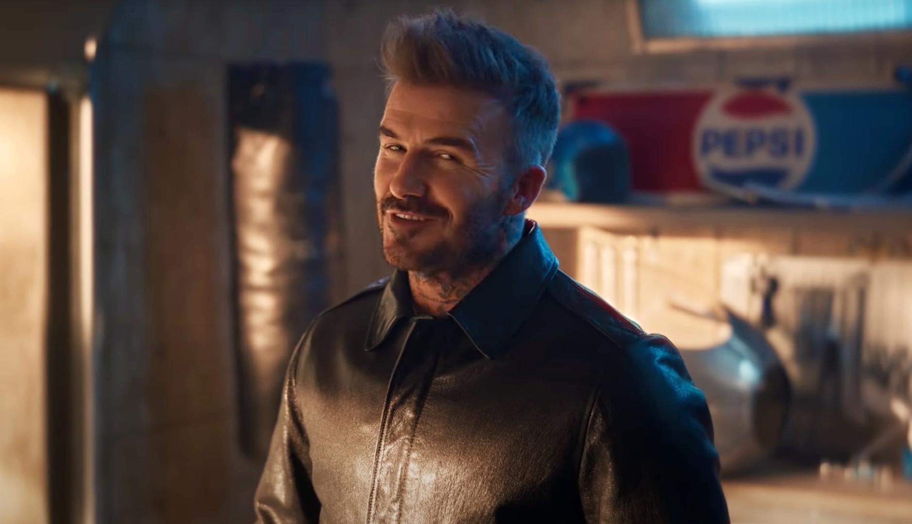 David Beckham sonriendo a cámara desde un garaje con un cartel de pepsi al fondo.