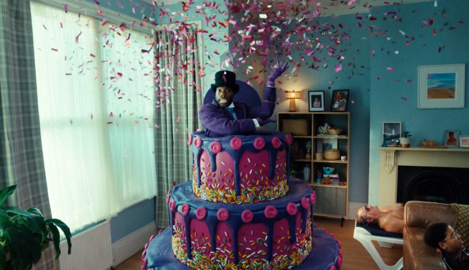 Un hombre saliendo de una tarta morada y rodeado de confeti en medio de un salón de casa