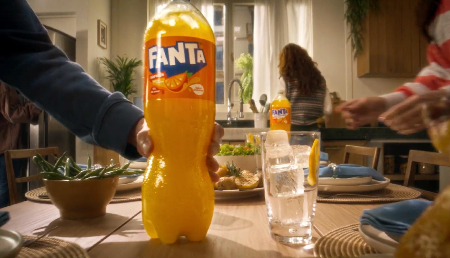 Una mano coloca una botella grande de Fanta sobre una mesa con la comida lista