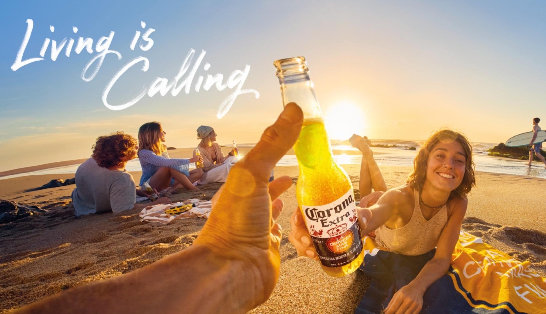 Una mano sostiene una Corona y brinda con una chica en un atardecer en la playa