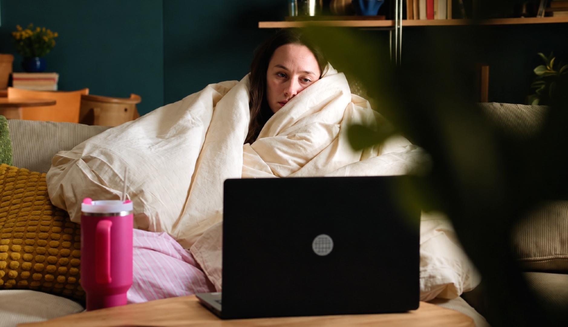 Una mujer con cara triste enrollada en su edredón en el sofá viendo una serie